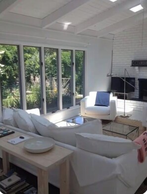 A modern living room with a white sofa, a coffee table, and a television. Large windows provide natural light and a view of greenery outside.