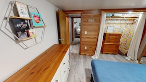 Ein Schlafzimmer mit einem hölzernen Schminktisch, einem Bett mit blauer Decke und einem an der Wand befestigten Regal mit Büchern.
