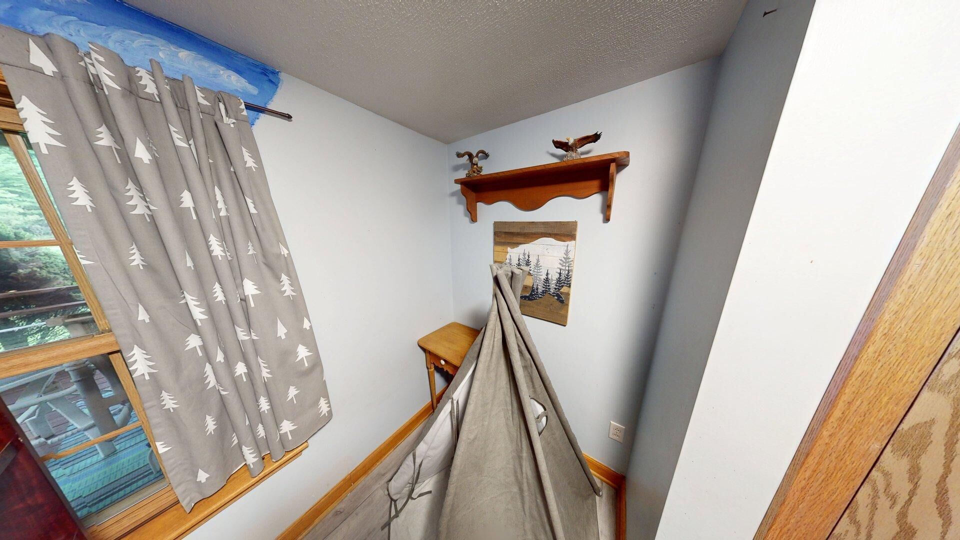 A room with a window, a curtain with a tree pattern, a small wooden table, a wall-mounted shelf with decorative items, and a framed picture.