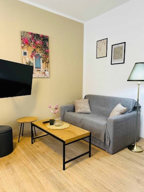 A living room with a grey sofa, a wooden coffee table, a small round table, a floor lamp, and a flat-screen TV.