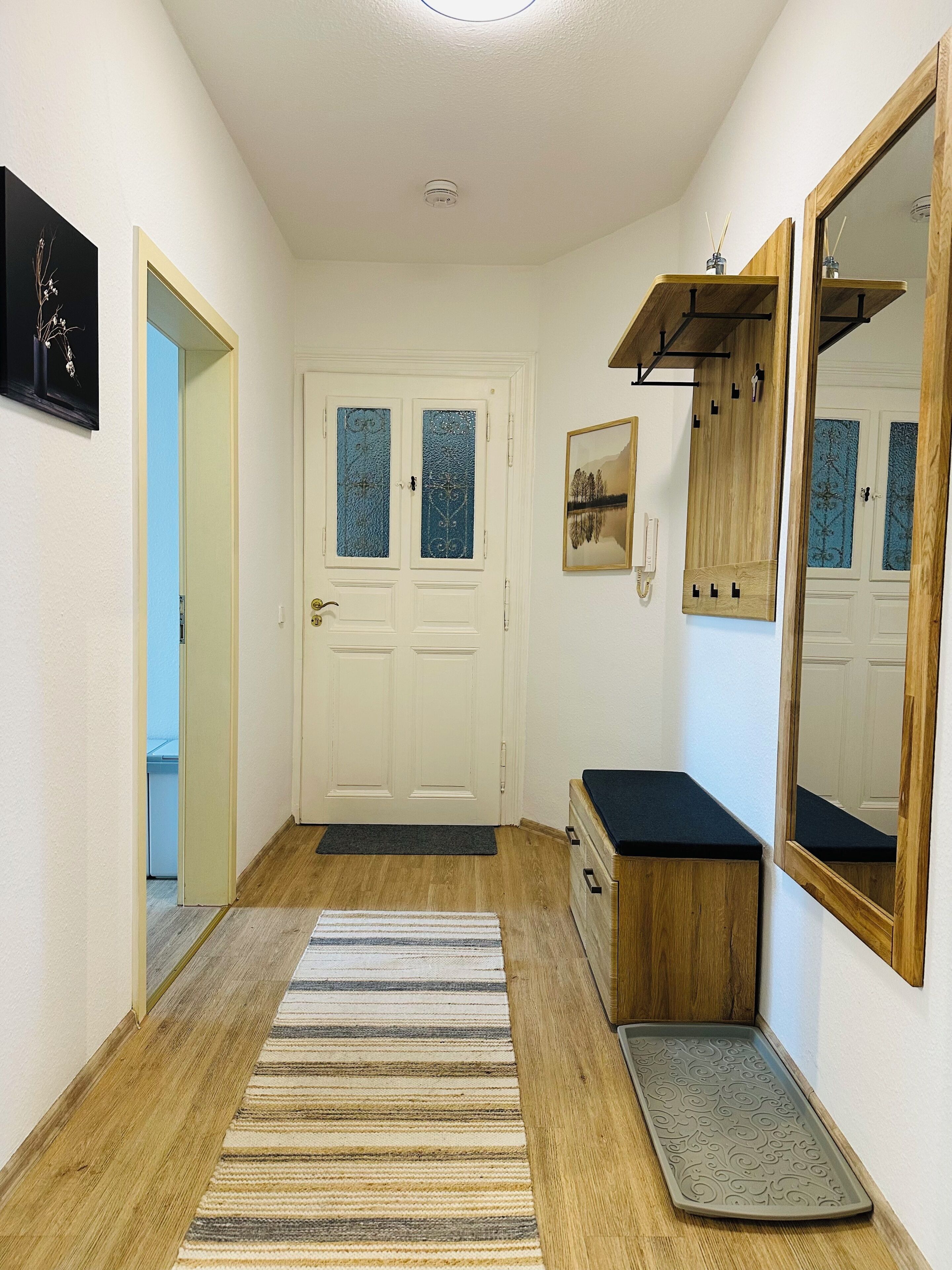 Un couloir doté d’un plancher en bois, d’un tapis à rayures, d’une porte avec des panneaux en verre dépoli, d’un miroir et d’une petite armoire en bois.