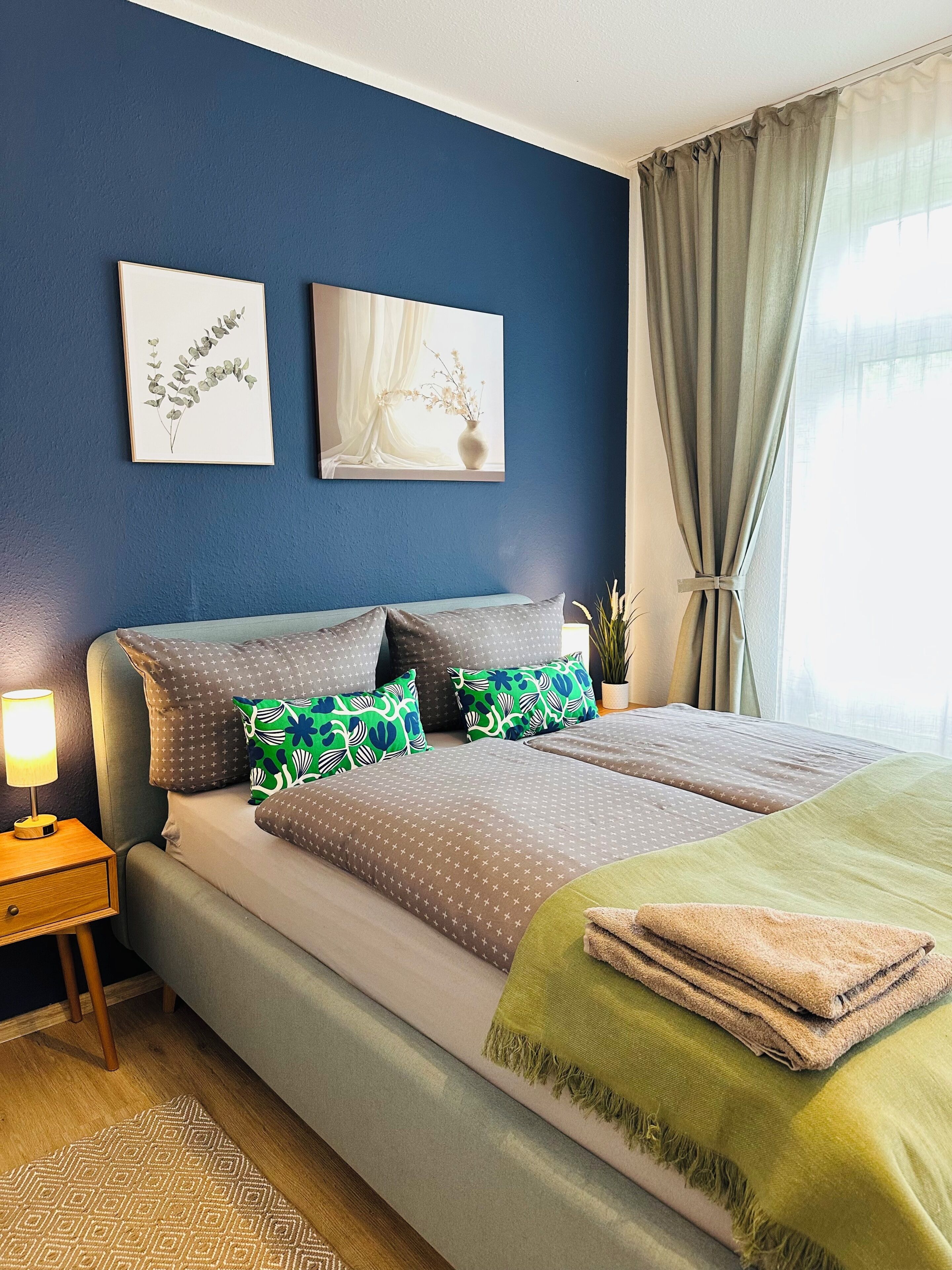 Un dormitorio con cama, almohadas, una mesita de noche, una lámpara y una obra de arte en la pared.