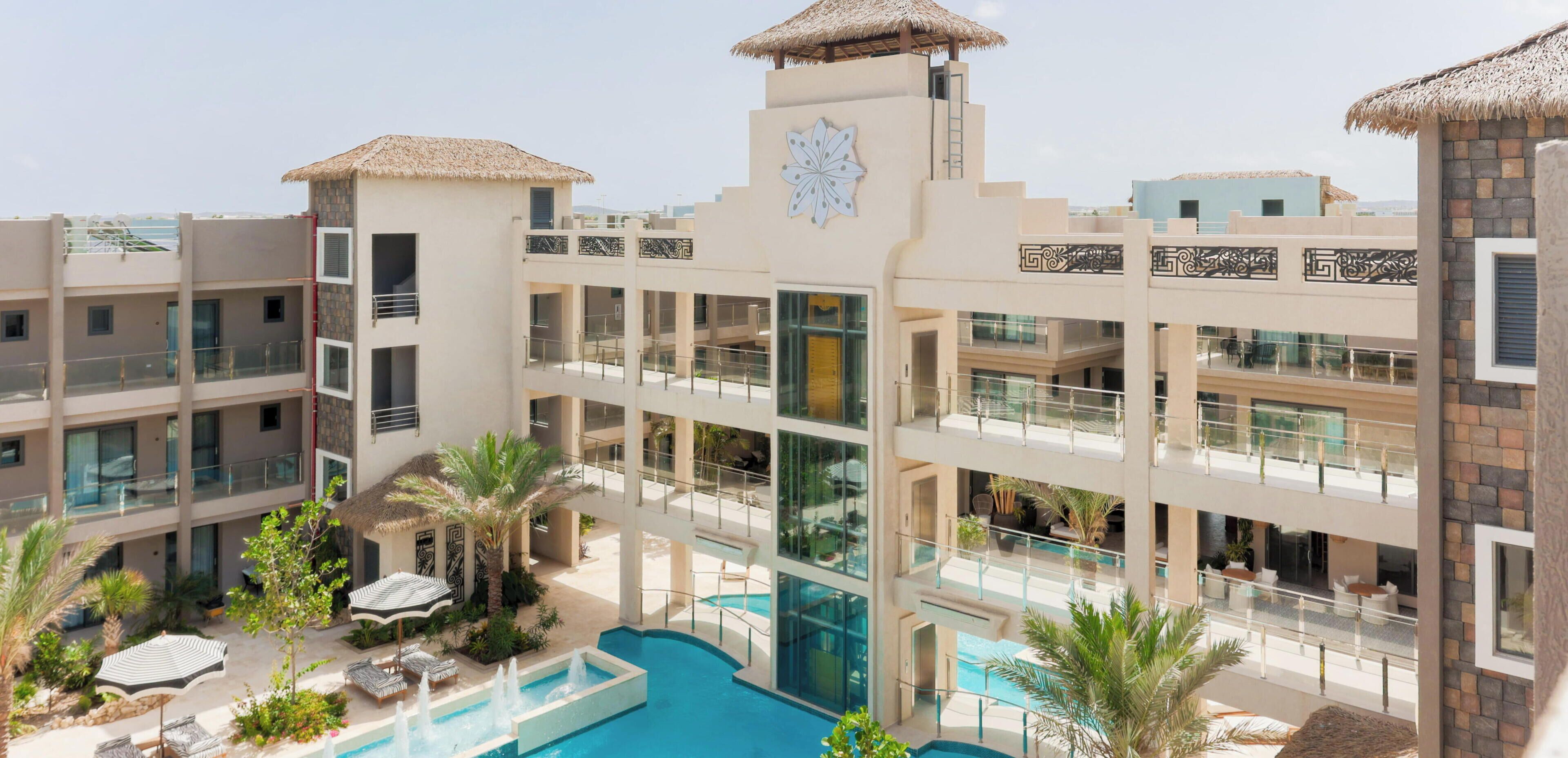 A multi-story hotel with a central courtyard featuring a swimming pool, lounge chairs, and palm trees.