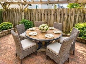 A round outdoor dining table with wicker chairs, surrounded by a wooden fence and greenery.