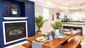 Área de refeições com mesa de madeira posta para seis, lareira e cozinha ao fundo.