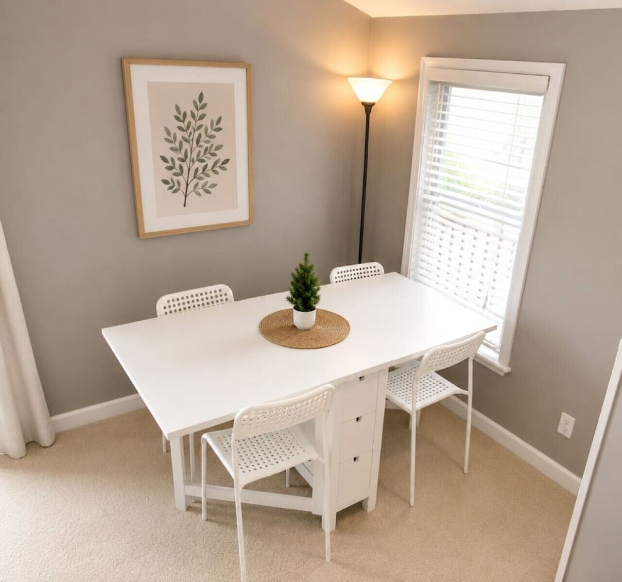 A white dining table with four chairs, a small potted plant, a framed leaf artwork, and a floor lamp.