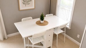 A white dining table with four chairs, a small potted plant, a framed leaf artwork, and a floor lamp.