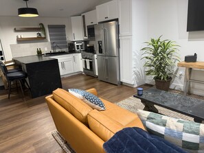 Eine moderne Küche mit weißen Schränken, Edelstahlgeräten, einer zentralen Kochinsel und einer Sitzecke mit Sofa und Couchtisch.