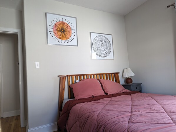 A bedroom with a wooden headboard, a pink bedspread, two framed artworks on the wall, and a bedside lamp.