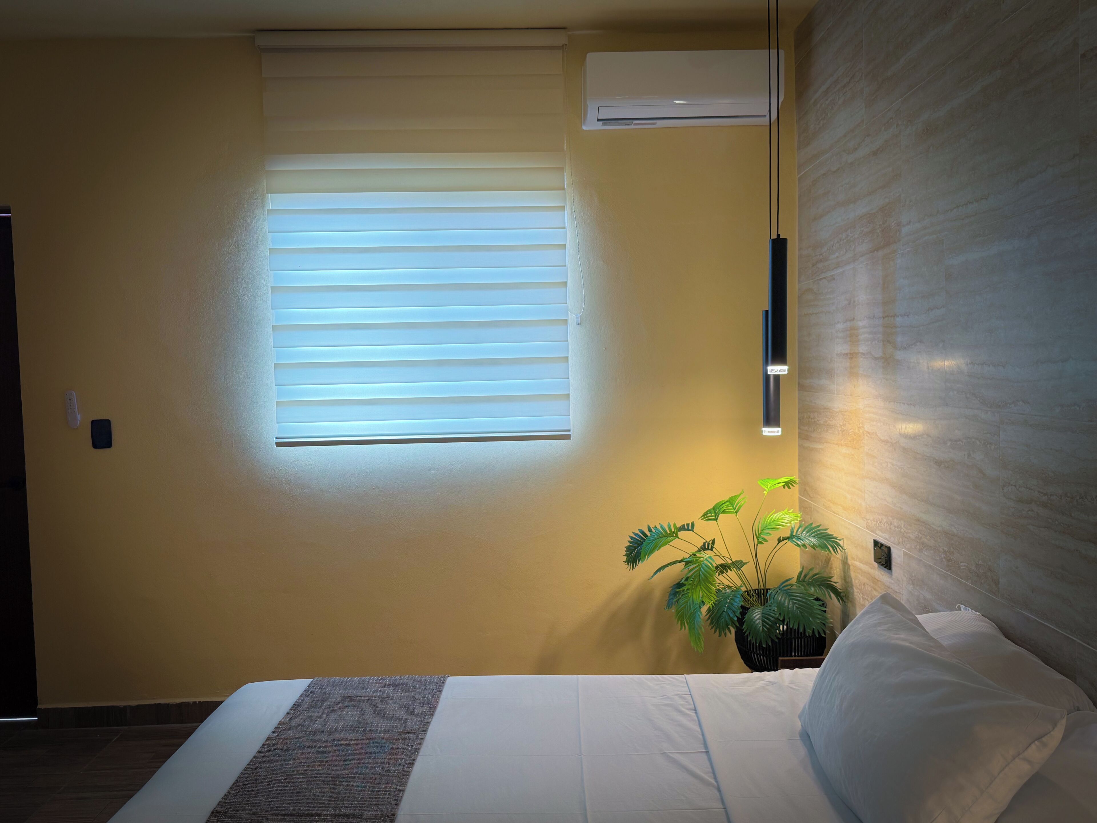 Une chambre d’hôtel avec un lit, une fenêtre avec des stores, un appareil de climatisation, un luminaire fixé au mur et une plante en pot.