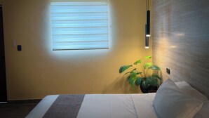 Hotelkamer met een bed, een raam met zonwering, een airconditioning, een wandlamp en een potplant.