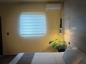 Et hotelværelse med seng, et vindue med persienner, en aircondition og en vægmonteret lampe samt en potteplante.