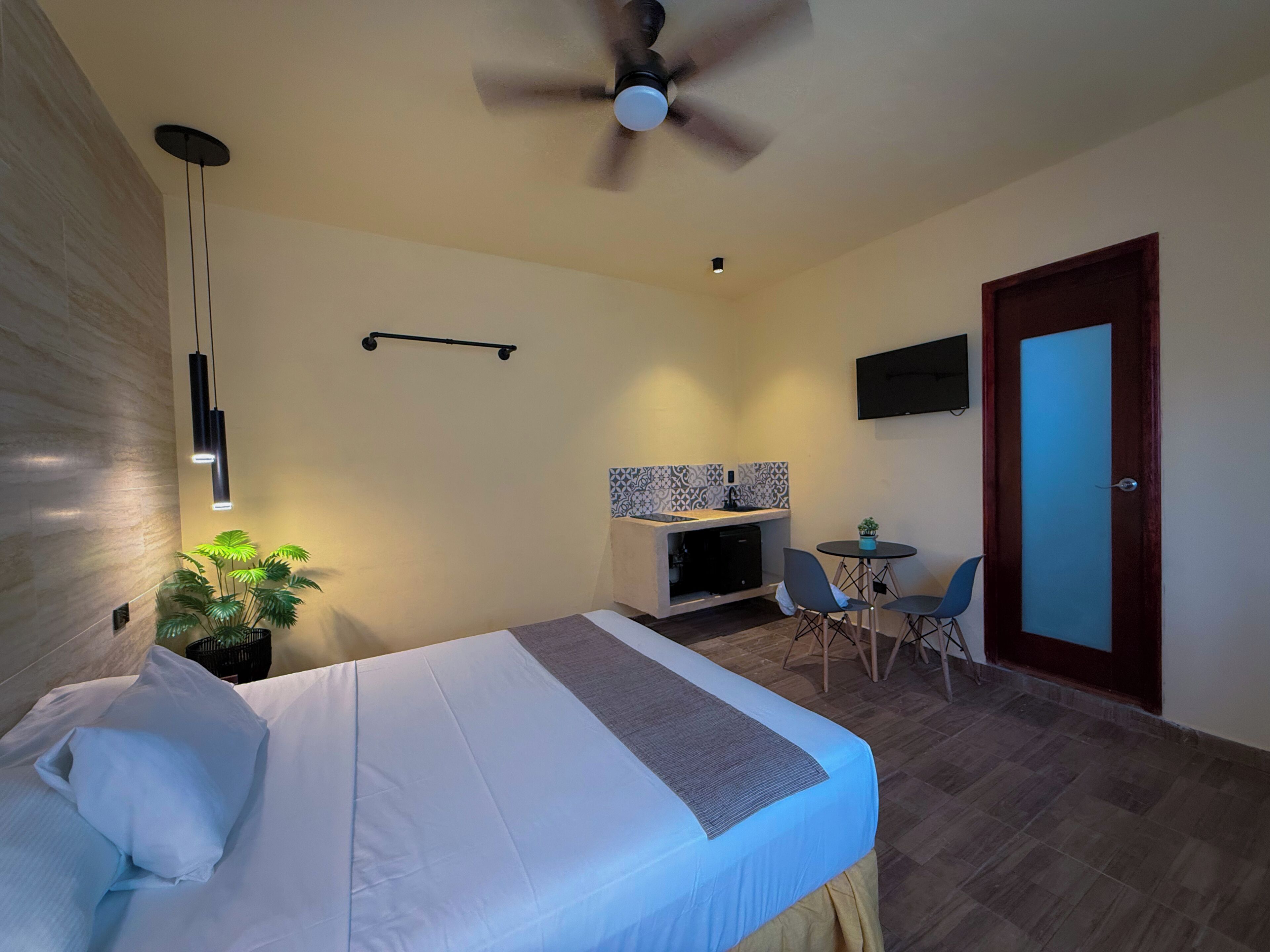 Een hotelkamer met een bed, een eettafel met stoelen, een plafondventilator, een aan de muur bevestigde televisie en een deur.