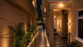 A nighttime corridor with a tiled floor, potted plants, and a staircase leading to a balcony with decorative railings.