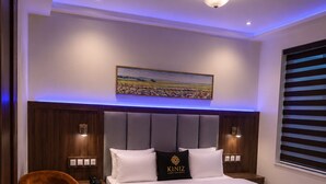 A modern bedroom with a large bed, a headboard with built-in lighting, a bedside table, a chair, and a desk with a remote control.