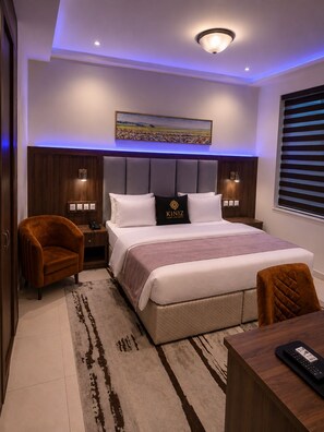 A modern bedroom with a large bed, a headboard with built-in lighting, a bedside table, a chair, and a desk with a remote control.