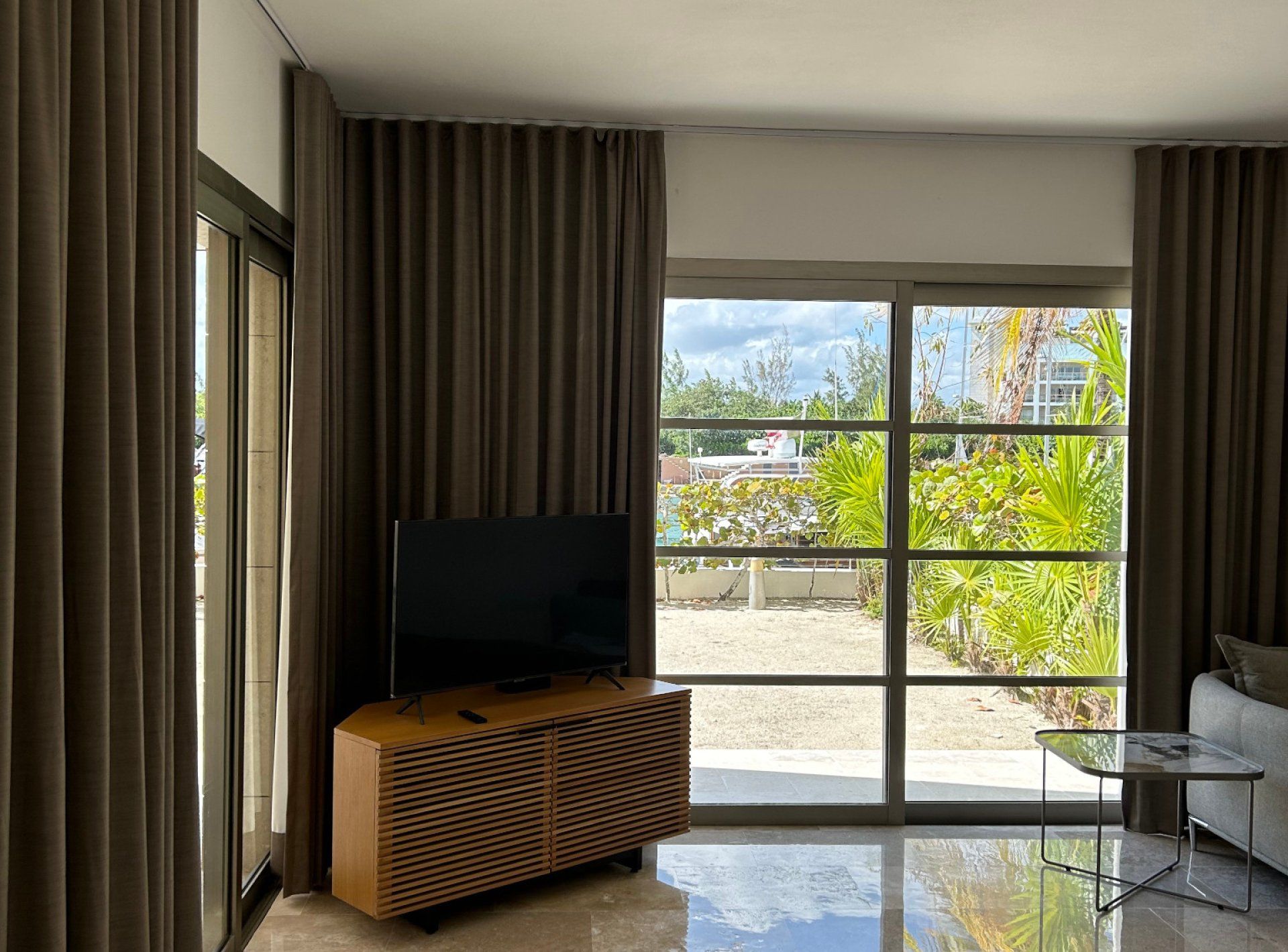 Uma sala de estar moderna com televisão de tela plana em um armário de madeira, janelas amplas com cortinas, e vista para áreas verdes e edifícios.
