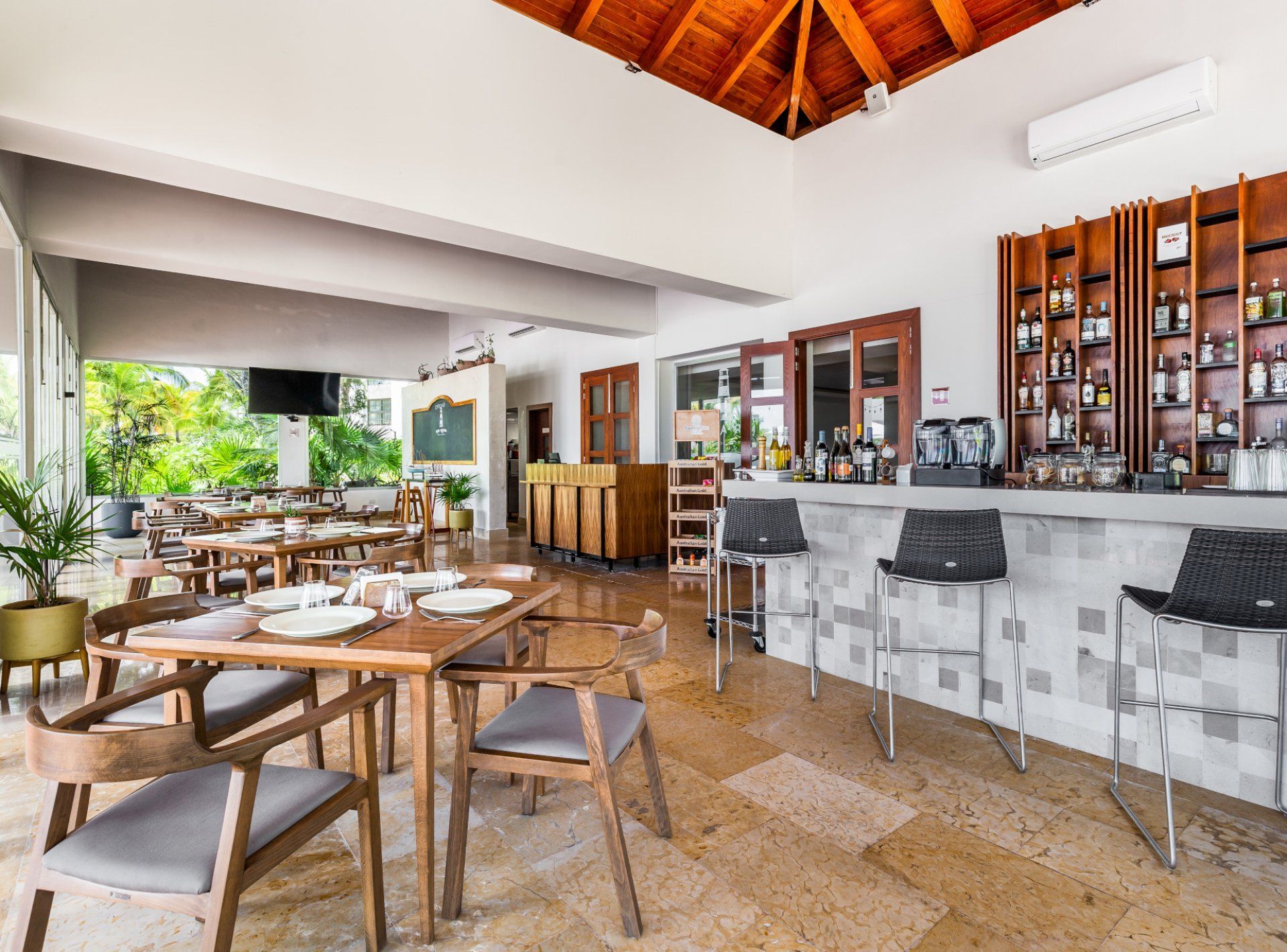 A spacious outdoor dining area with wooden tables and chairs, a bar with a tiled backsplash, and a view of lush greenery outside.