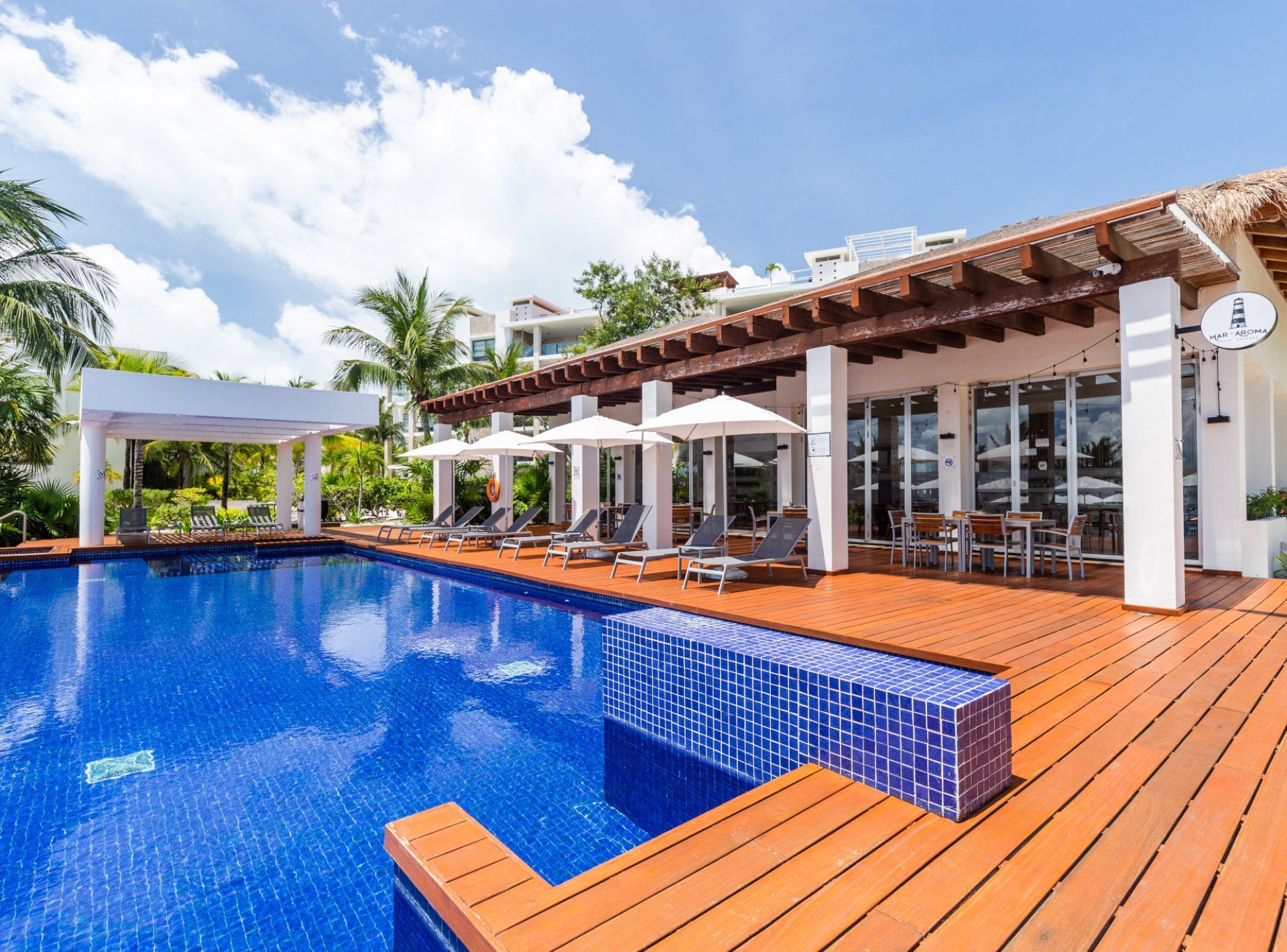 A swimming pool with lounge chairs and umbrellas, surrounded by a wooden deck and a covered area with tables and chairs.