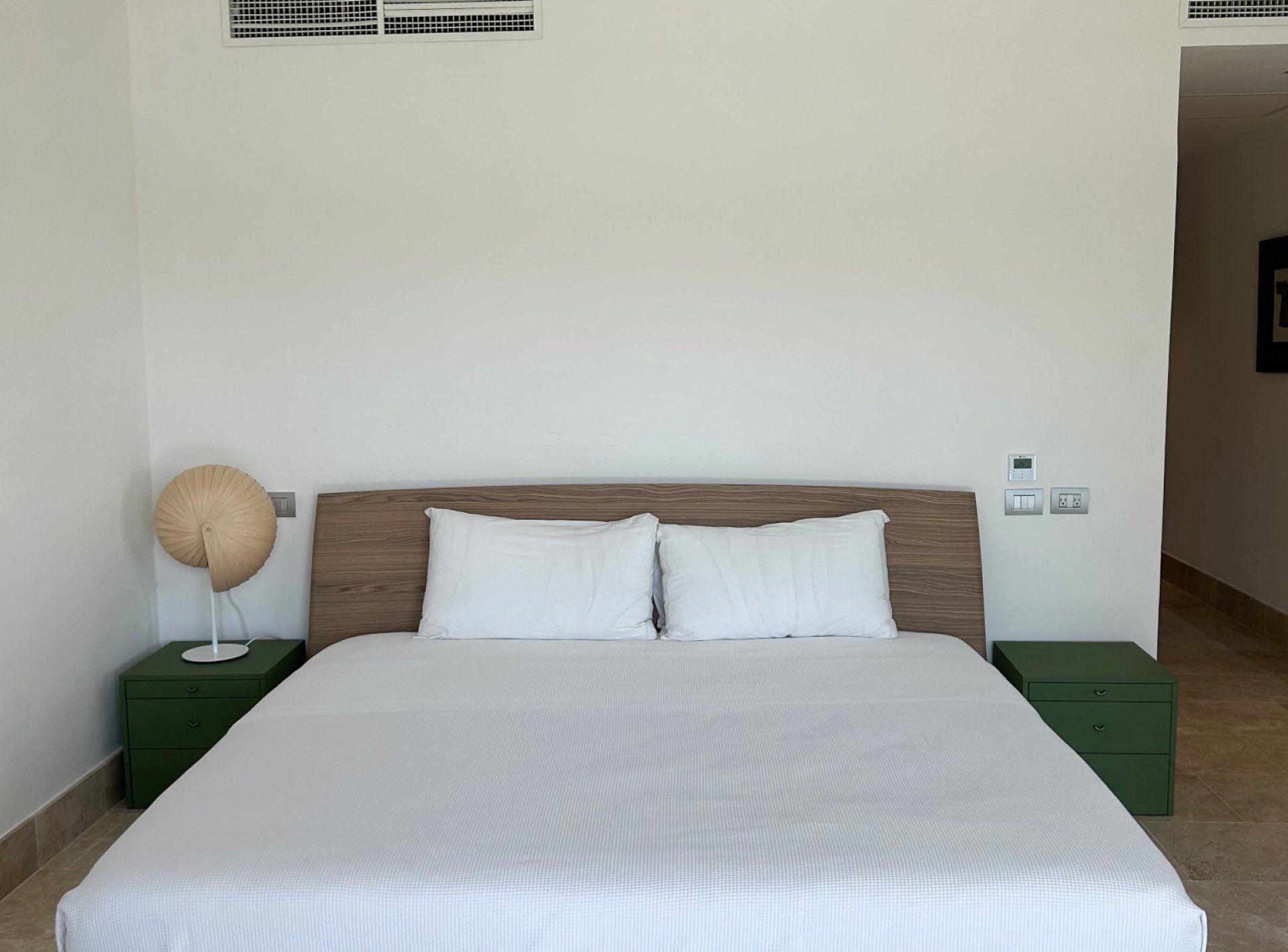 Una habitación de hotel con una cama grande, dos mesitas de noche, un respiradero en la pared y una mesita de noche verde con una lámpara.