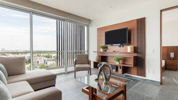 Ein modernes Wohnzimmer mit einem Glastisch, einer Couch, einem an einer Holzplatte montierten Fernseher und Blick auf die Stadt durch große Fenster.