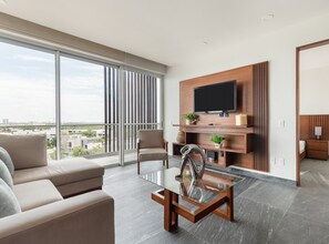 Un salon moderne avec une table basse en verre, un canapé, un téléviseur fixé sur un panneau en bois et une vue sur la ville grâce à de grandes fenêtres.