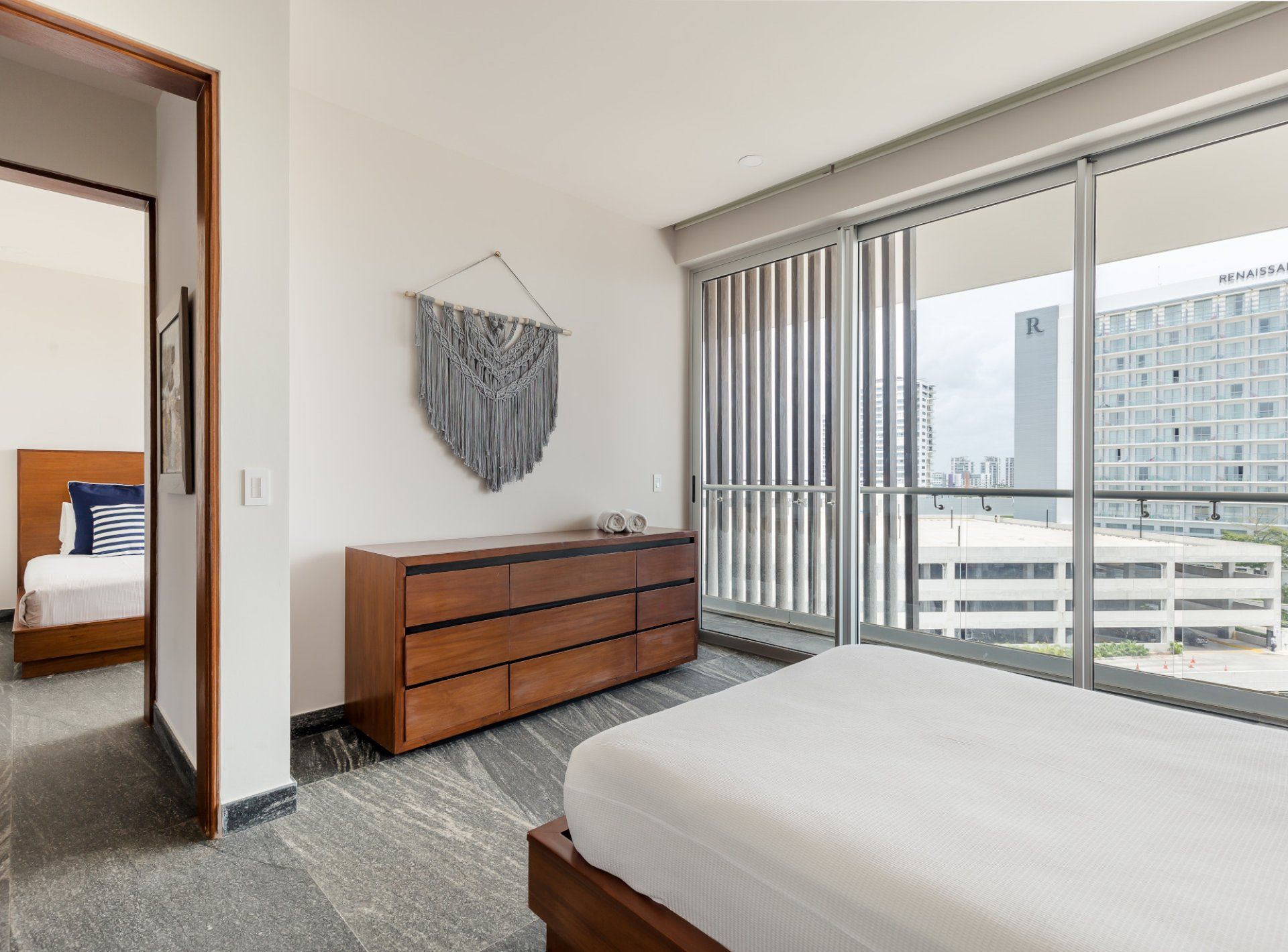 Ein modernes Hotelzimmer mit einem großen Bett, einer Holzkommode, einem Wandbehang aus Makramee und Blick auf eine Stadtlandschaft durch raumhohe Fenster.