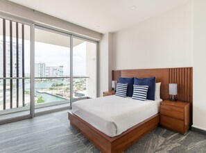 Une chambre d’hôtel moderne dotée d’une grande porte-fenêtre coulissante donnant sur un balcon, d’un lit bien fait avec des oreillers à rayures, d’une table de chevet en bois et d’une vue sur le paysage urbain et l’eau.