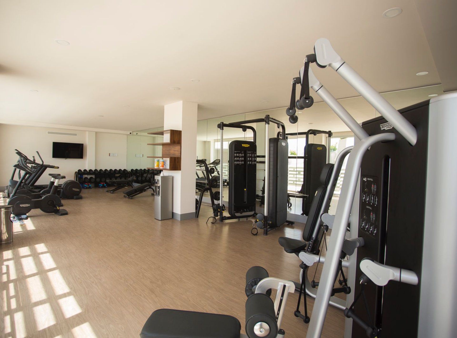 A gym with various exercise equipment, including treadmills and weight machines, and a mirror wall.