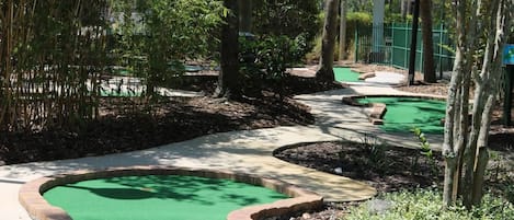 A miniature golf course with multiple green putting areas, surrounded by trees and a covered pavilion.