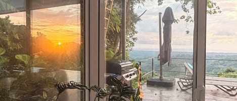Un intérieur moderne offrant une vue sur un coucher de soleil sur l’eau, avec un barbecue et un espace salon extérieur.