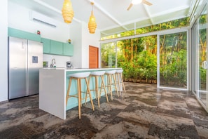 A modern kitchen with a central island, bar stools, and a view of lush greenery through large windows. - Your tropical retreat overlooking Corcovado, featuring a private pool and lush natural surroundings. (Puerto Cortes)