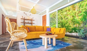 A room with a yellow sofa, a wicker chair, a round wooden table, and a large window with a view of greenery. - Your tropical retreat overlooking Corcovado, featuring a private pool and lush natural surroundings. (Puerto Cortes)