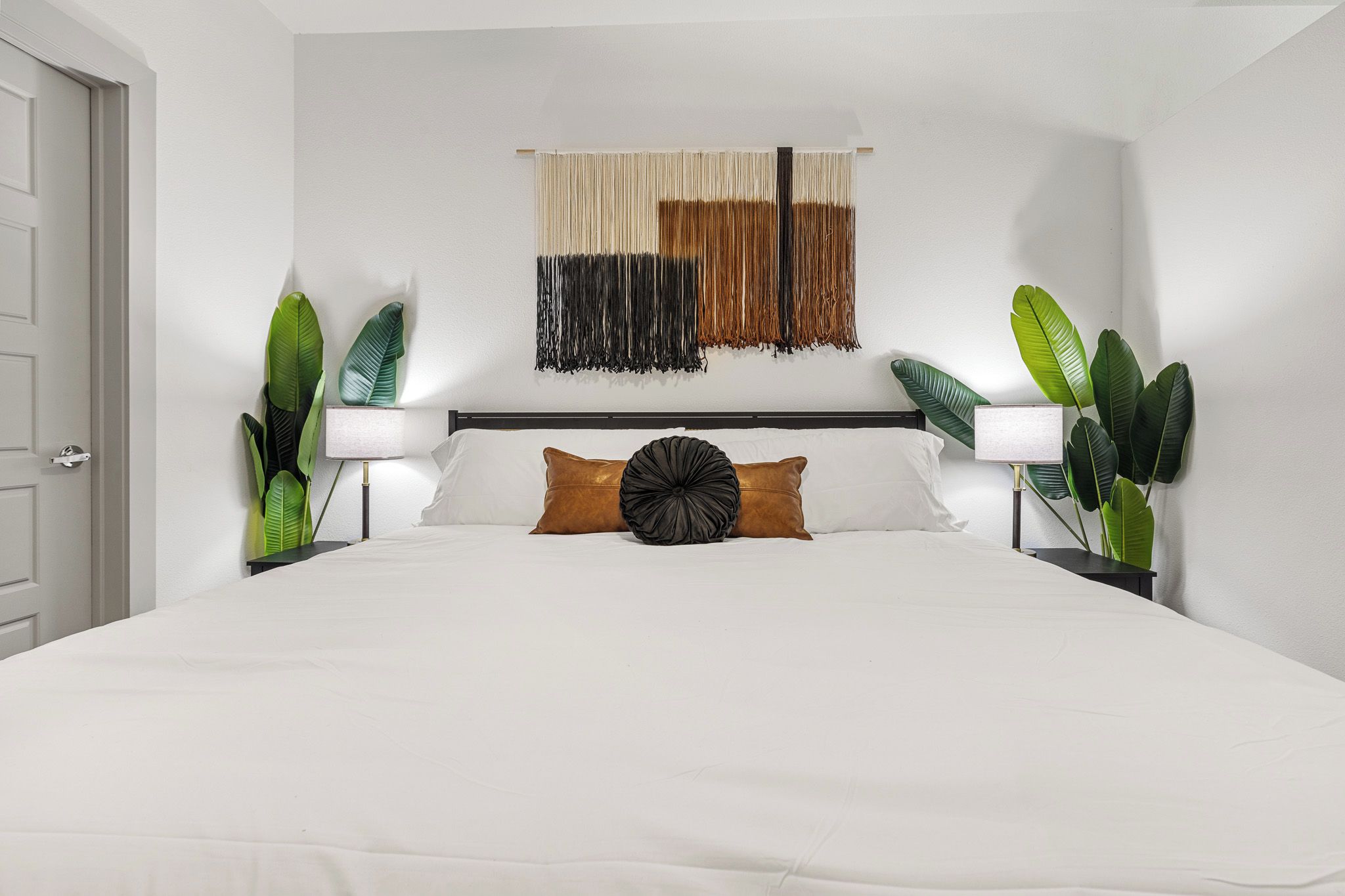 A hotel room with a large bed, two bedside lamps, a wall-mounted tapestry, and potted plants.