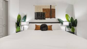 A hotel room with a large bed, two bedside lamps, a wall-mounted tapestry, and potted plants.