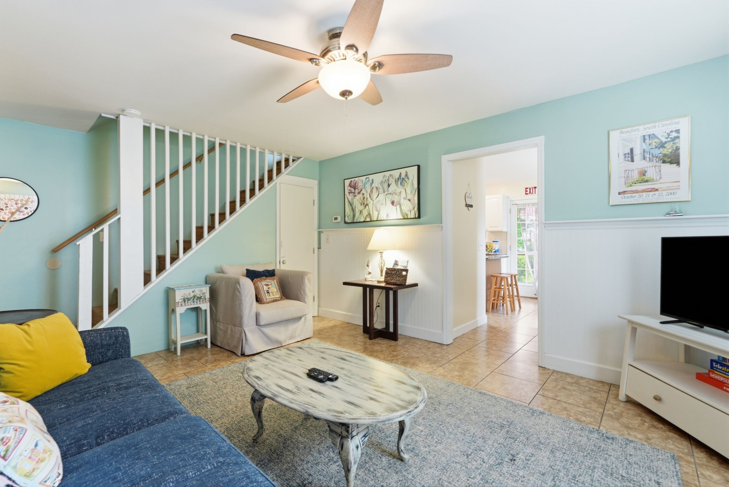 A living room with a blue sofa, a coffee table, a ceiling fan, a staircase, and a door leading to another room.