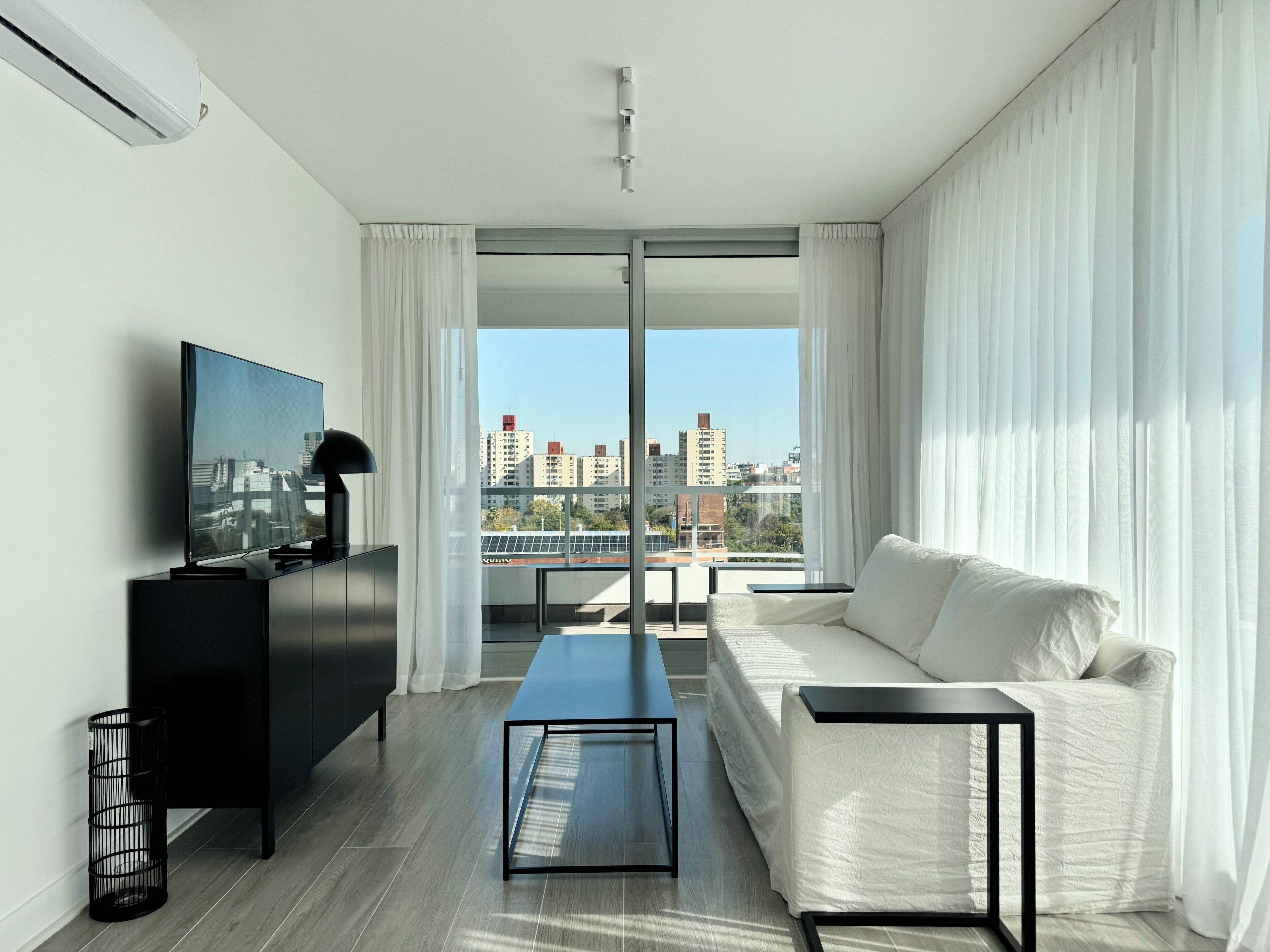 A modern living room with a flat-screen TV, a sofa, a coffee table, and a view of a cityscape through large windows.