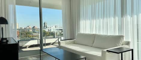 A modern living room with a white sofa, a black coffee table, and a view of the city through large windows.