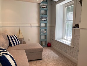 A room with a beige sofa, a blue and white striped pillow, a small sailboat decoration, a red exercise ball, and a window with a view of greenery.