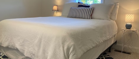 Una cama bien hecha con cubrecama y almohadas blancas, un tapete estampado, una ventana con persianas y dos lámparas de noche.