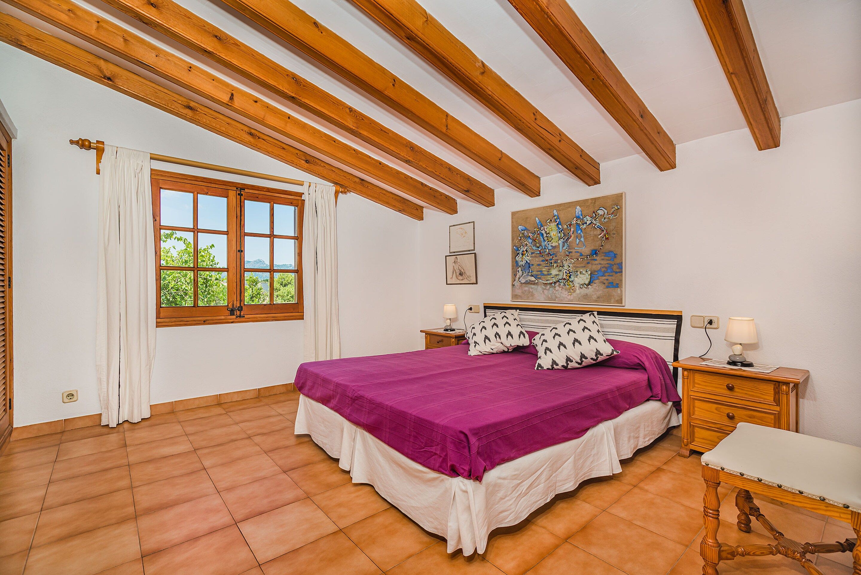 A bedroom with a bed, a nightstand, a window with curtains, a painting on the wall, and wooden beams on the ceiling.
