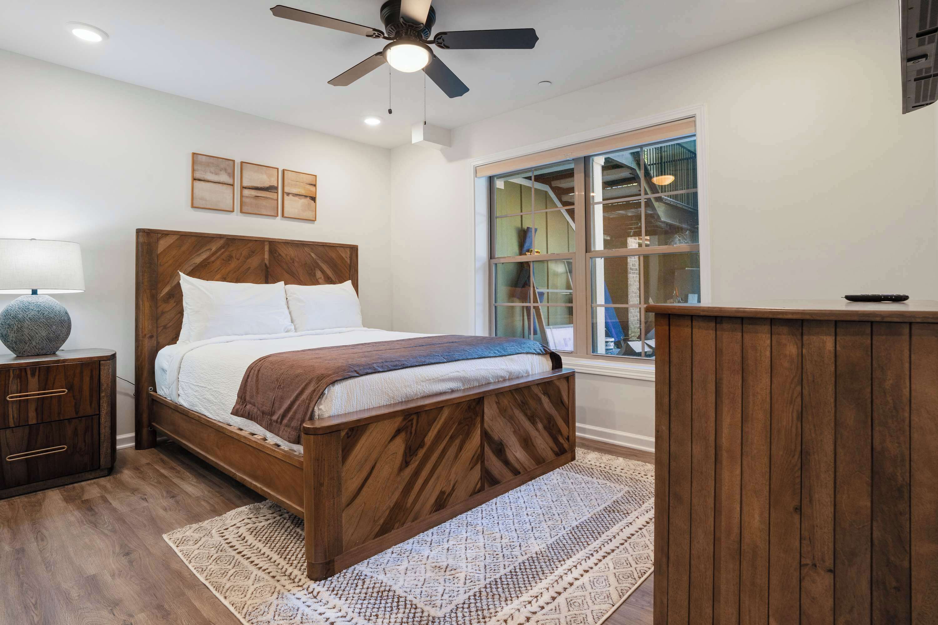A bedroom with a wooden bed, a nightstand, a lamp, a window with a view, and a ceiling fan.