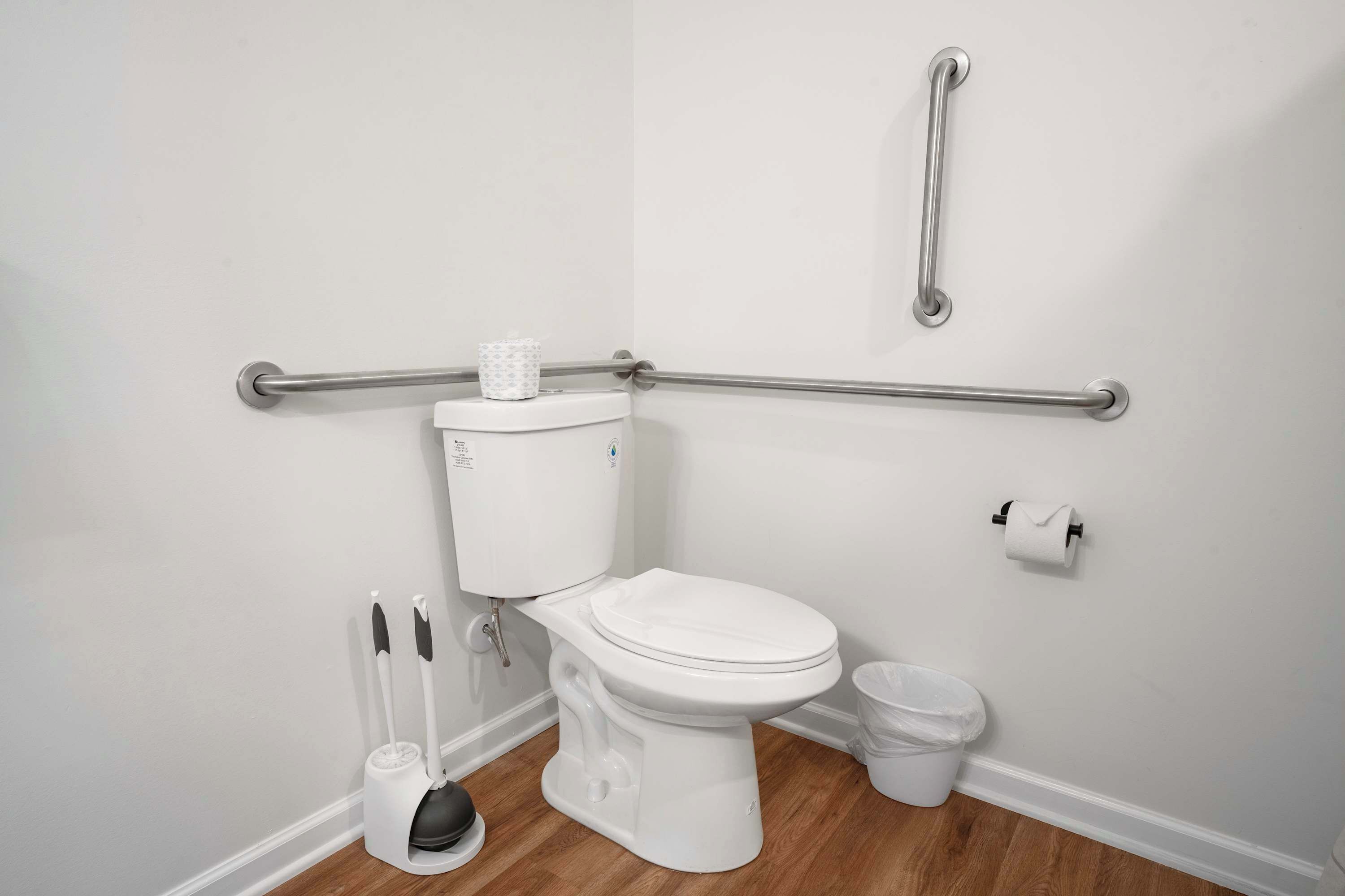 A white toilet with a bidet attachment, a grab bar, a toilet paper holder, and a trash bin.