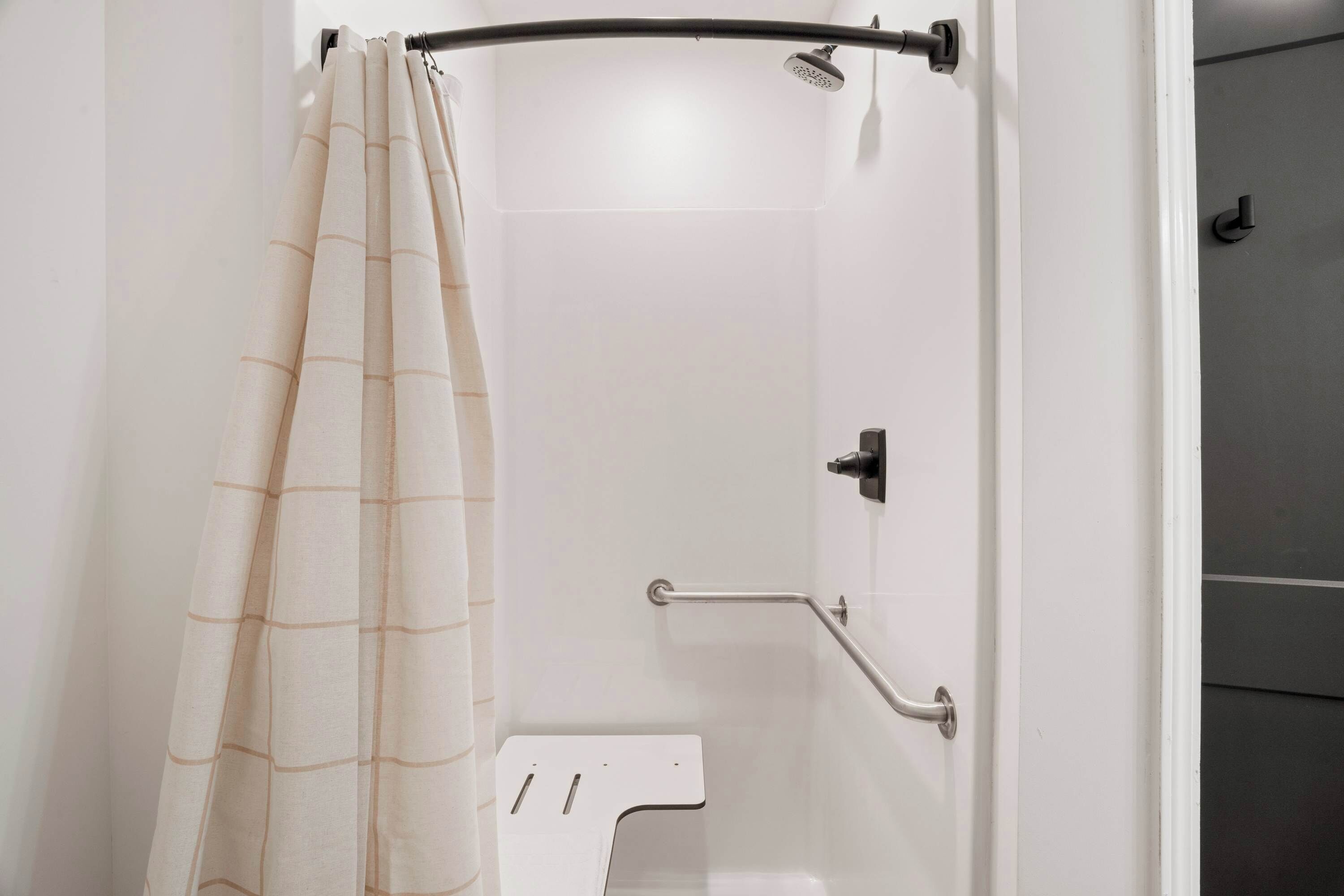 Un área de ducha con pared de azulejos blancos, cabezal de ducha, barra de apoyo y cortina.