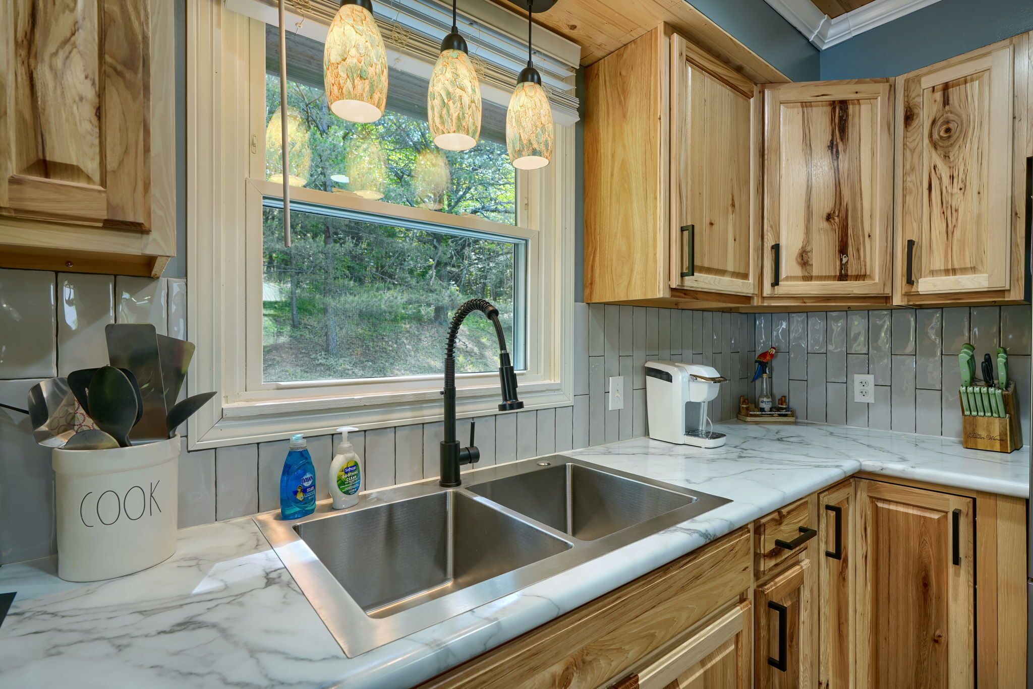 A kitchen with a double sink, wooden cabinets, a window with a view of greenery, and pendant lights.