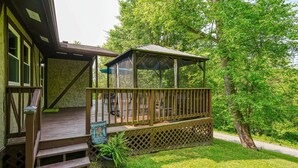 A wooden deck with a covered seating area, a sign indicating a hide-and-seek champion, and a lush green lawn. - Hideaway Pines Cabin (Logan)