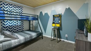 A bunk bed with a patterned headboard, a Pac-Man arcade machine, a television, and a plant on a wooden cabinet. - Hideaway Pines Cabin (Logan)