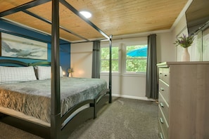 A four-poster bed with a quilted mattress, a painting on the wall, a window with a view of greenery, and a dresser with drawers. - Hideaway Pines Cabin (Logan)