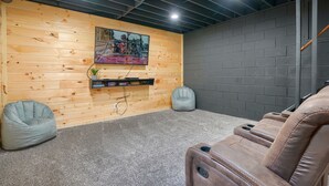 A room with a wooden wall, a television mounted on it, a grey bean bag chair, a brown reclining chair, and a grey carpet. - Hideaway Pines Cabin (Logan)