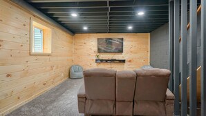 A room with a wooden wall, a ceiling with visible pipes, a mounted television, and a brown leather sofa. - Hideaway Pines Cabin (Logan)